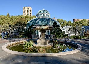 The image shows a botanical garden conservatory with a dome, surrounded by greenery and featuring a central fountain with a circular pond.