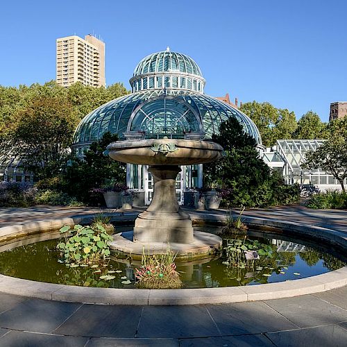 The image shows a botanical garden conservatory with a dome, surrounded by greenery and featuring a central fountain with a circular pond.