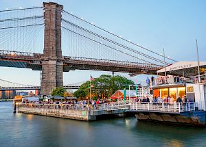 The image shows a waterfront scene with a bridge in the background. There's a dock area with people dining and a building labeled 