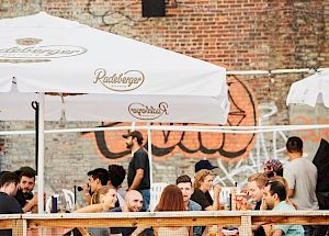 People dining outdoors under large, branded umbrellas with a brick wall and graffiti in the background.