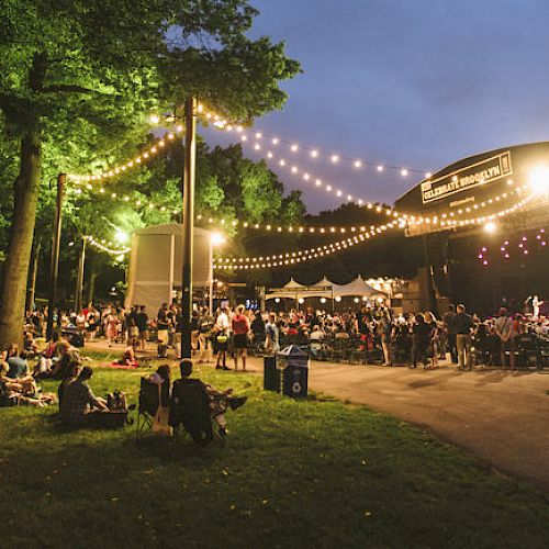 A night-time outdoor concert with string lights, people sitting on the grass, and a stage with performers under the illuminated canopy.