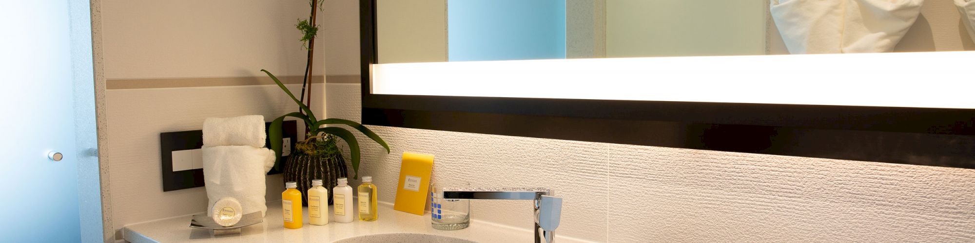 A modern bathroom with a sink, mirror, toiletries, towels, a potted plant, and a toilet. The lighting is bright, and the decor is minimalistic.