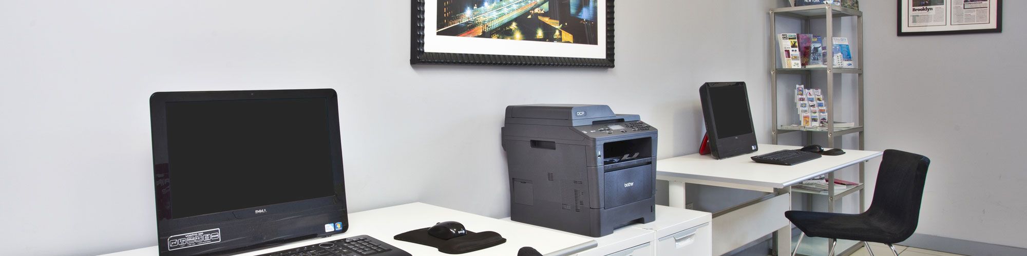 The image shows a small office space with computers, a printer, chairs, framed pictures, and a shelf with books and magazines.