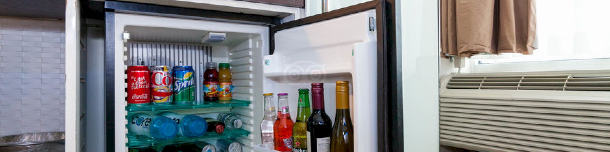 An open mini fridge in a hotel room is stocked with various beverages, including soft drinks, water, and wine bottles.