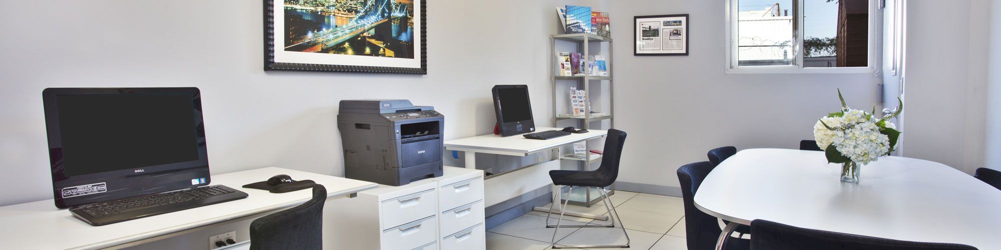 The image shows an office with desks, computers, a printer, chairs, a round table with flowers, wall art, a bookshelf, and a window in the background.