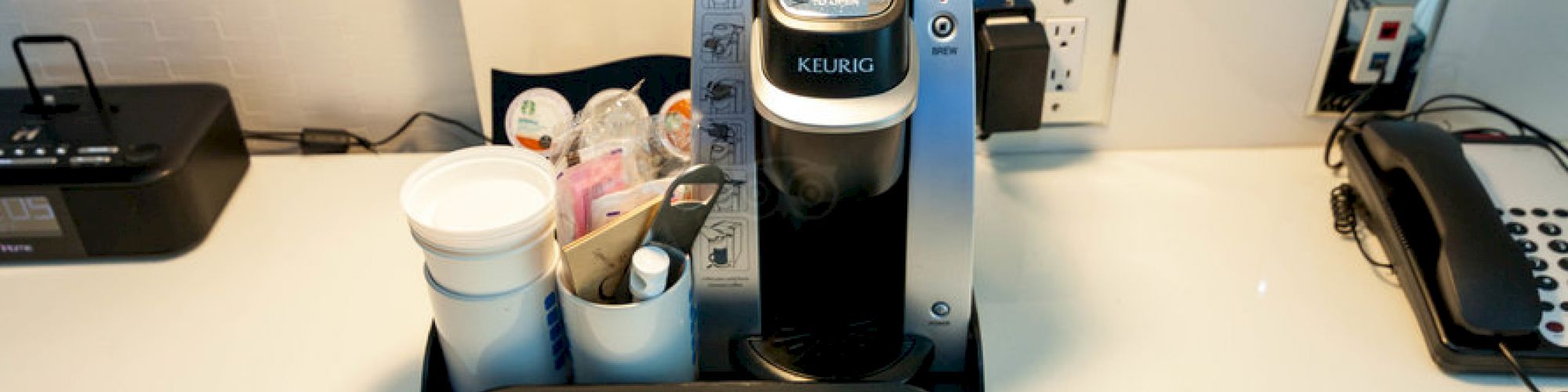 The image shows a Keurig coffee machine on a counter with cups, coffee pods, and sugar packets arranged in a tray, next to a phone.