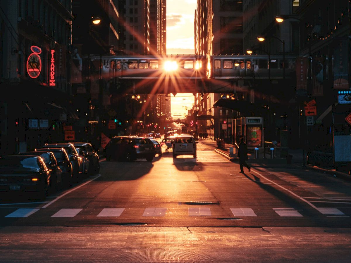 A sunlit urban street features cars, pedestrians, shops, and a train on a bridge overhead, with a striking sunset and illuminated signs in the background.