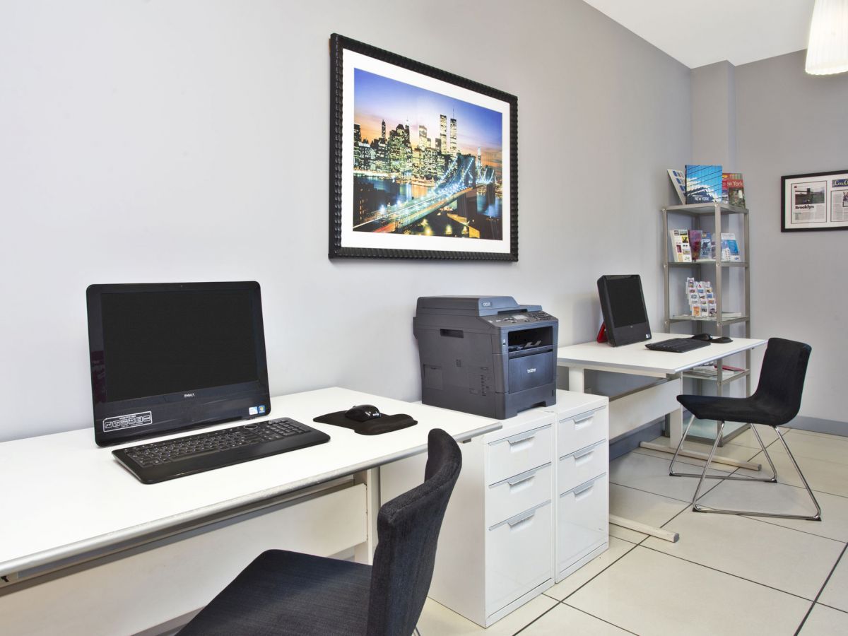 The image shows a small office area with two desks, computers, a printer, chairs, and a wall-mounted picture of a cityscape.