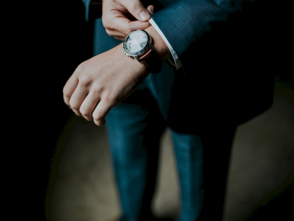 A person in a suit adjusts their watch, focusing on their hands and wrists, with brown shoes visible below.