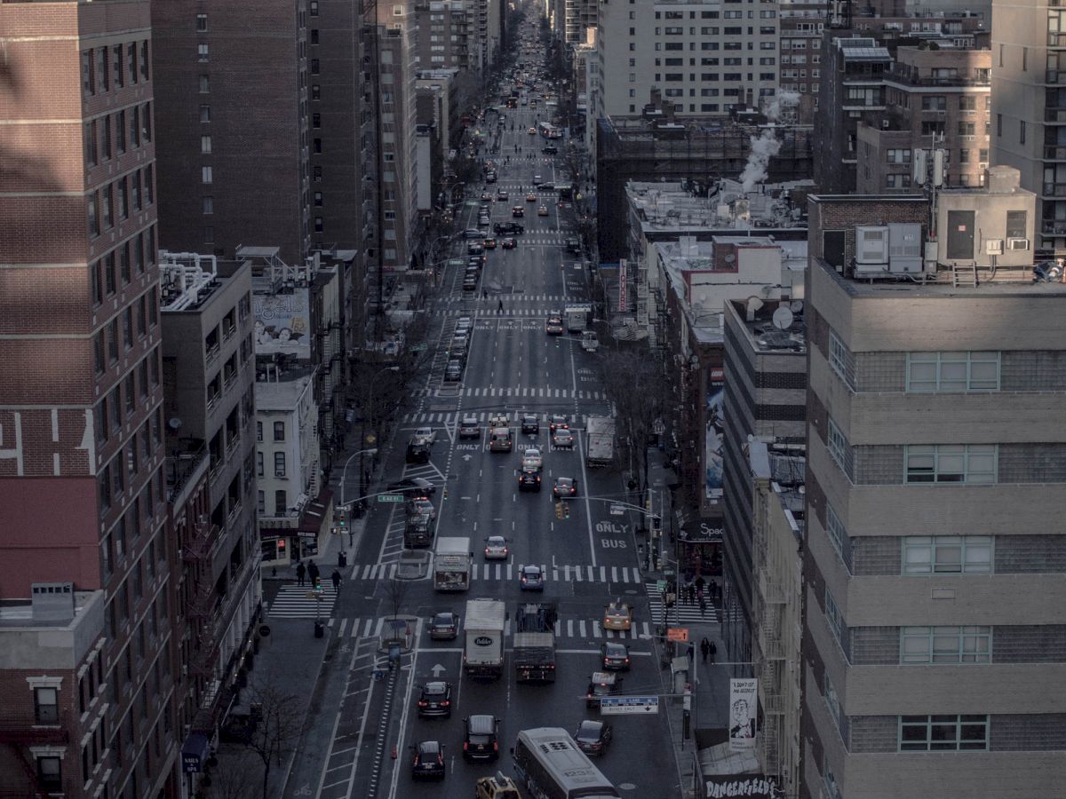 The image shows a busy city street with tall buildings lining both sides, numerous vehicles, and people going about their day.