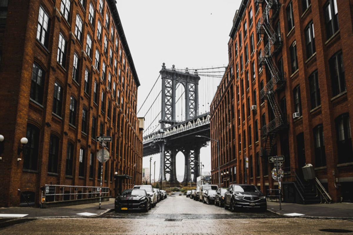 The image shows a street view framed by brick buildings, leading to a bridge structure in the distance on an overcast day.