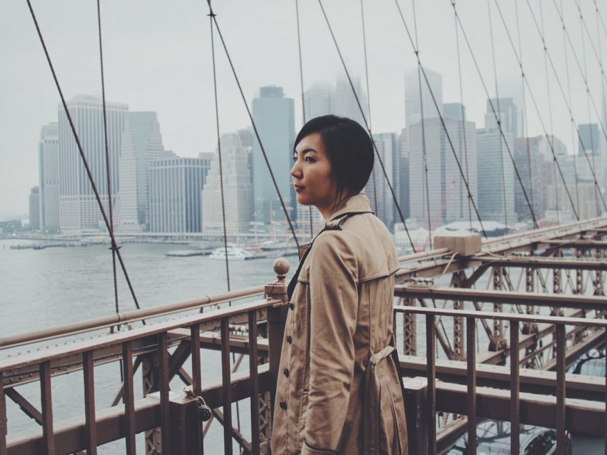 A person in a trench coat stands on a bridge with a city skyline in the background on a foggy day, gazing into the distance, deep in thought.