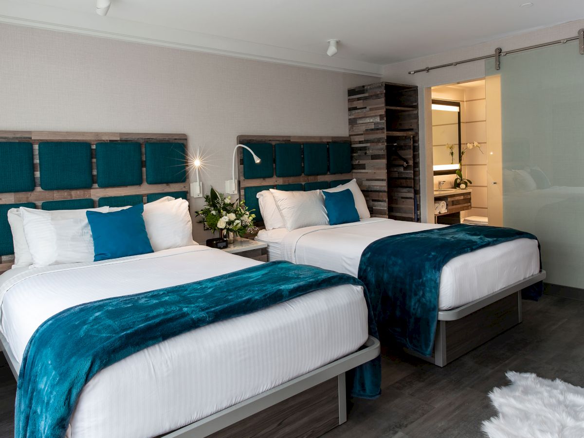 The image shows a modern hotel room with two beds, blue accents, white linens, a sliding glass bathroom door, and wood decor on the walls.