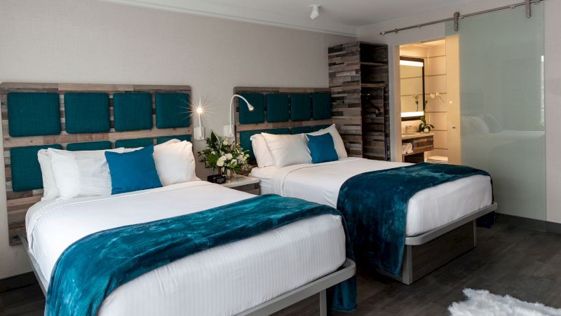 A hotel room with two beds, wooden headboards with teal cushions, a nightstand with flowers, and a glass door to the bathroom.