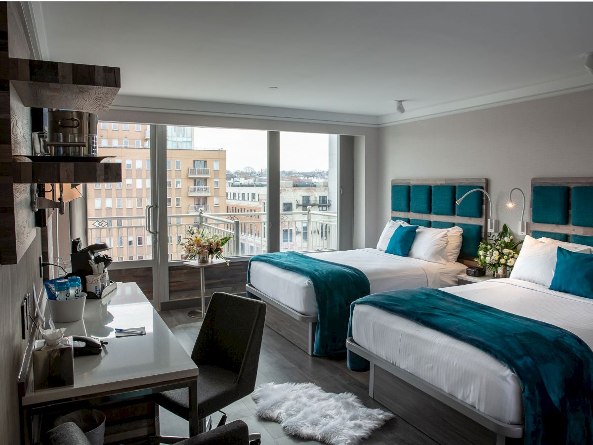 A modern hotel room with two beds, teal accents, a desk, a chair, a fluffy rug, and large windows overlooking city buildings and flowers.