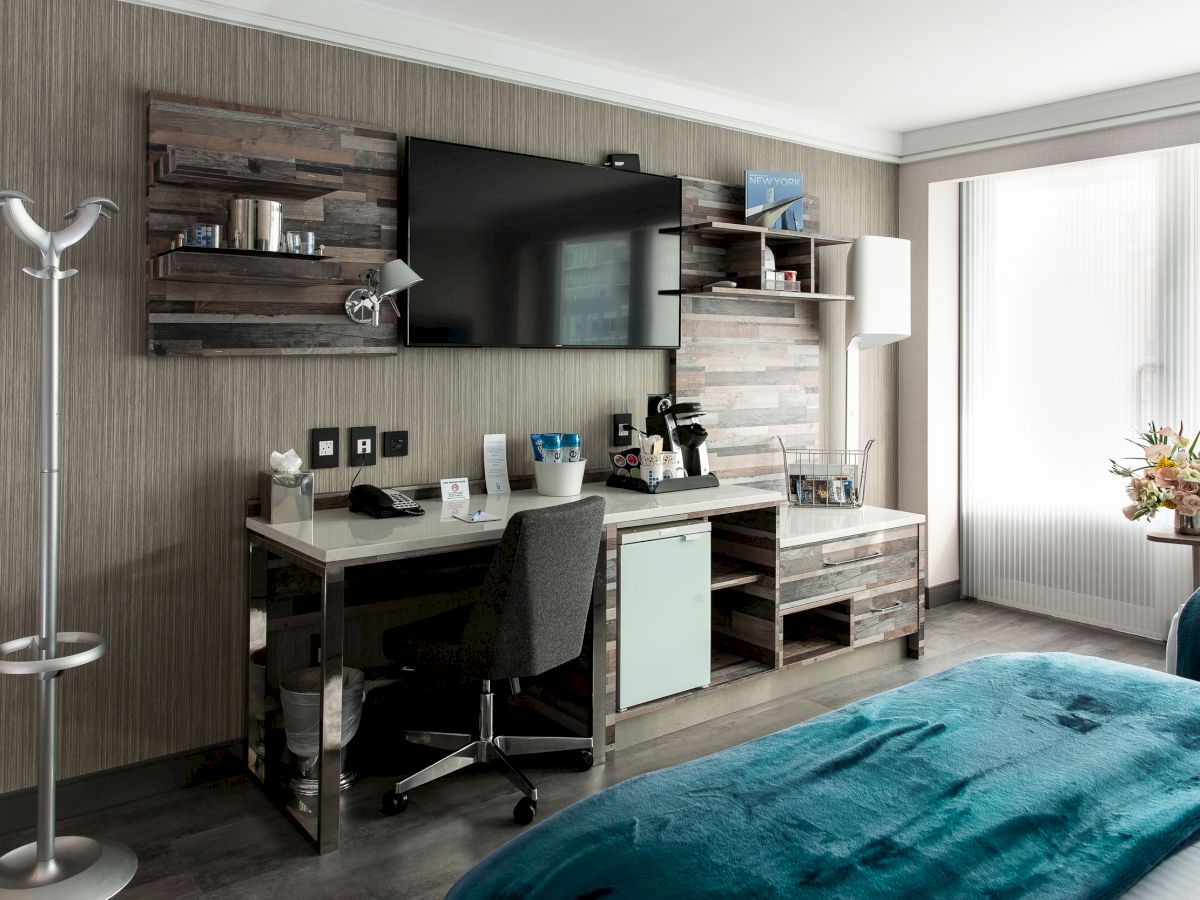 A modern hotel room with a desk setup, TV, shelves, and a flower vase on the cabinet by the window, with blue bedding visible on the beds.