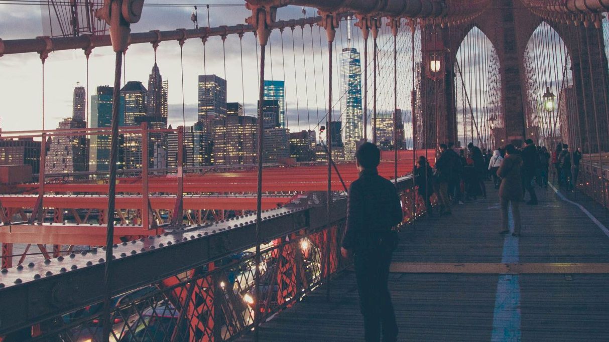 People walking on a bridge at dusk, with a city skyline in the background illuminating the scene.