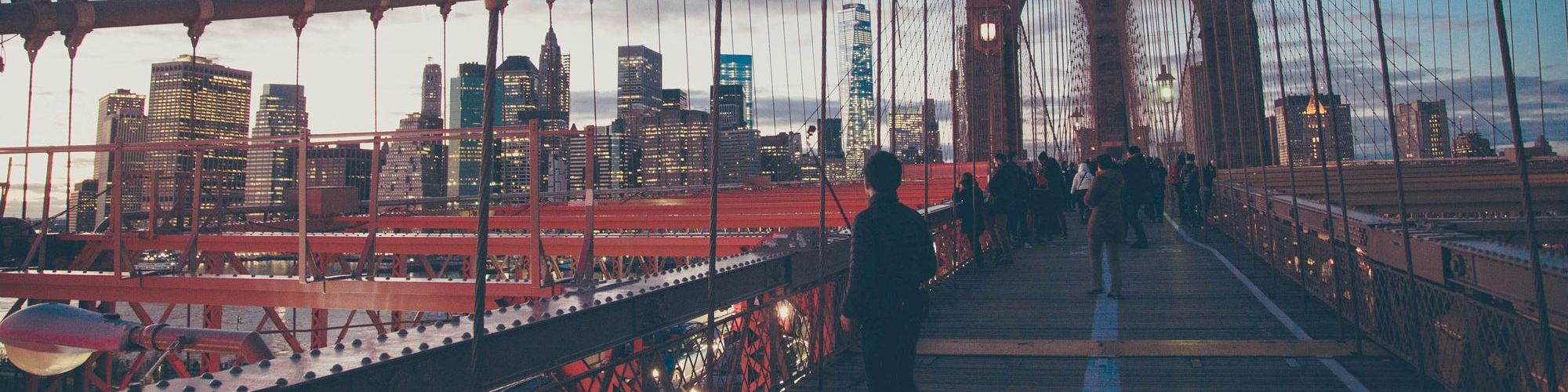 People walking on a bridge at dusk, with a city skyline in the background illuminating the scene.