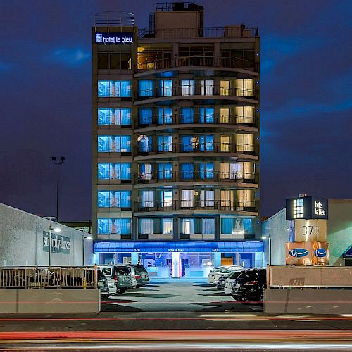 The image shows a multi-story building with blue lighting, situated at night. Vehicles are parked nearby, and light trails are visible on the road.