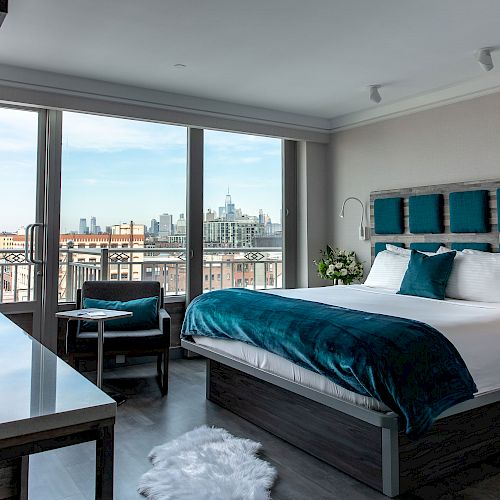 A modern hotel room with a large bed, desk, chair, and city view through large windows. The decor features blue accents and contemporary design.