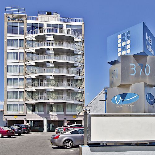 The image shows a multi-story building with balconies, a parking lot with cars, and a large sign in the foreground displaying 