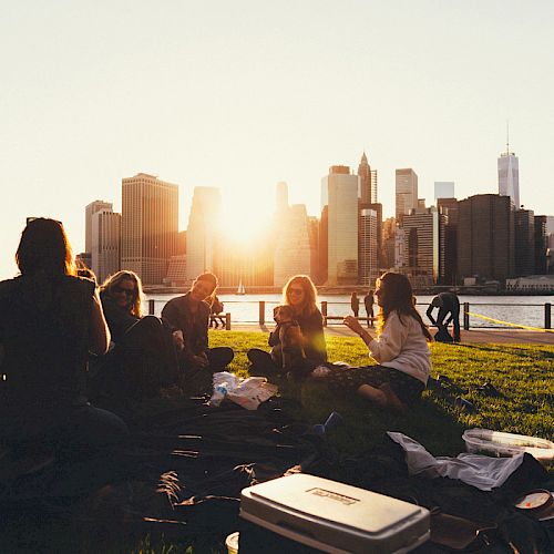 A group of people having a picnic on a grassy area with a city skyline in the background during sunset.