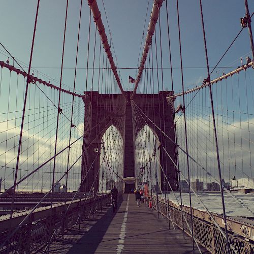 A view of the Brooklyn Bridge in New York City with cables, walkway, and city skyline in the background, under a partly cloudy sky.