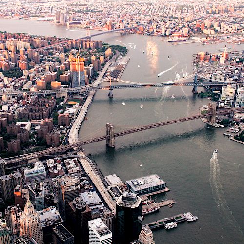 Aerial view of New York City featuring multiple bridges spanning the East River, with densely packed buildings and waterways dotted with boats.