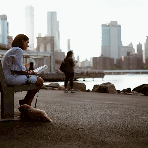 A person sits on a bench reading with a dog beside them, overlooking a city skyline near the water as another person walks by.