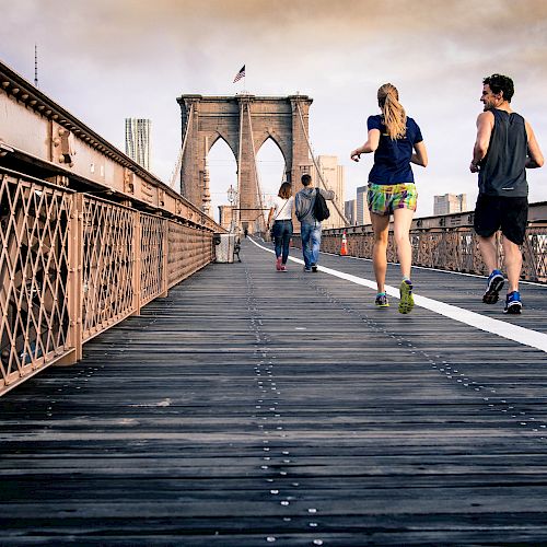 People are walking and running on a designated path of the Brooklyn Bridge, with the New York City skyline in the background.