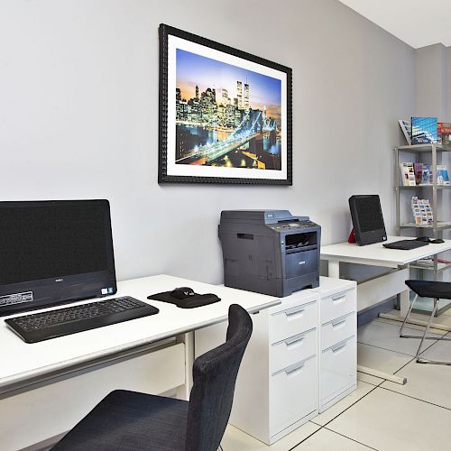 The image shows an office space with desks, computers, a printer, office chairs, shelves with books, and framed artwork on the wall.