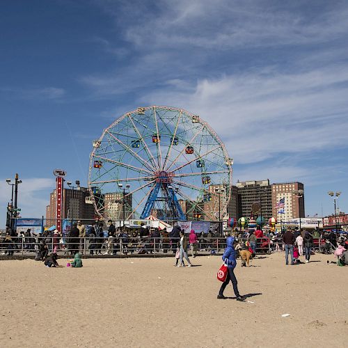 A beach scene featuring a Ferris wheel in the background, with people walking on sand and various buildings and structures visible.