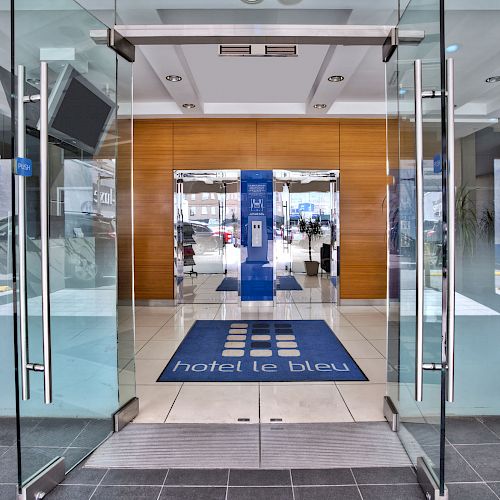 The image shows the entrance of a modern hotel with glass doors, a blue welcome mat reading 