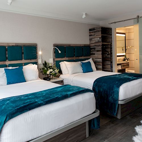 The image shows a hotel room with two double beds. Each bed has blue and white bedding. There's a sliding door leading to a bathroom.