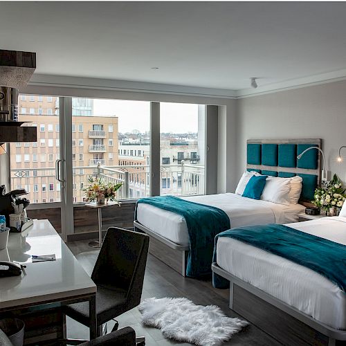 A modern hotel room with two beds, teal accents, a desk, chair, and city view through large windows. The room also has a bookshelf and coffee machine.