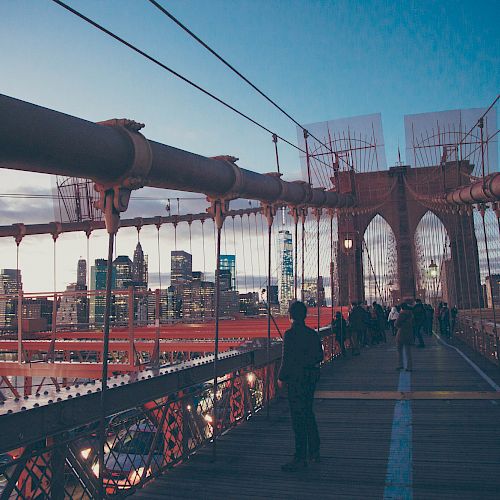 The image shows people walking on the Brooklyn Bridge with the New York City skyline visible in the background during dusk.