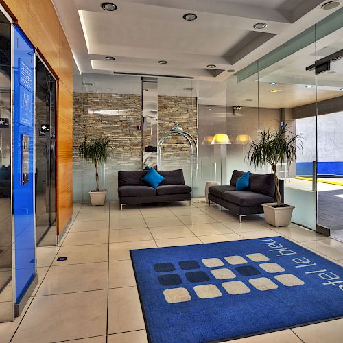 The image shows a modern hotel lobby with blue accents, including a blue mat, couches with blue pillows, and a blue sign by the elevators.