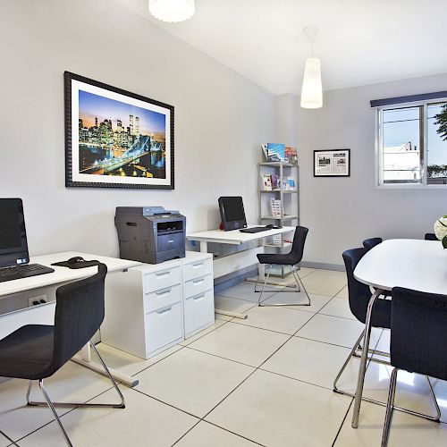 A modern office with desks, chairs, computers, a printer, a table with flowers, shelves, and a wall picture.