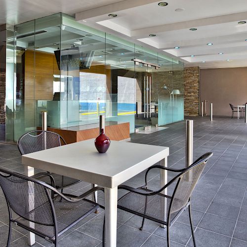 A modern indoor seating area with a table and two chairs; a vase sits on the table. The background includes a glass wall and another seating area.