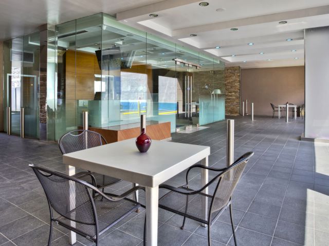 A modern lobby with a white table and black chairs, surrounded by glass walls and a tiled floor.