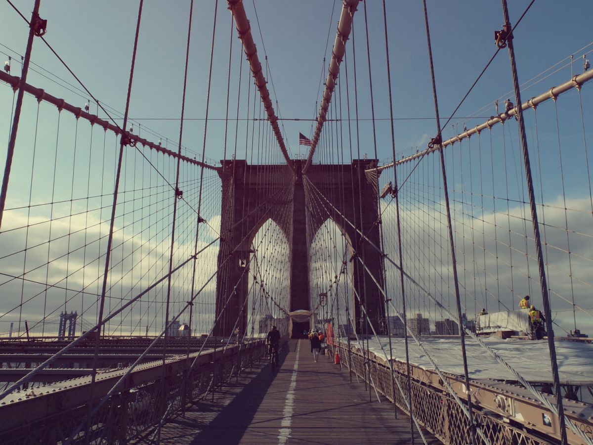 This image shows a pedestrian walkway on a large suspension bridge with cables and towers, likely the Brooklyn Bridge, with people walking along it.