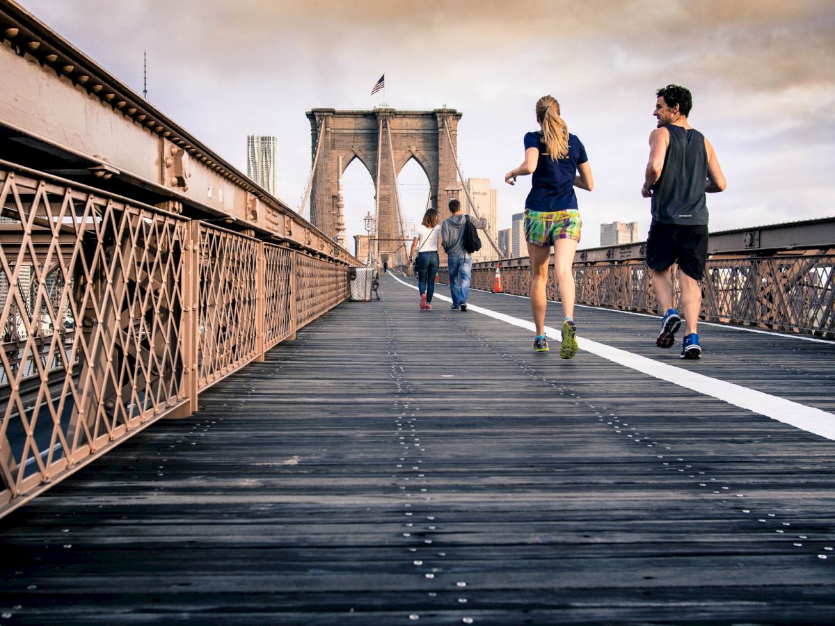 Three people are running and one person is walking on a wooden bridge with an iconic architectural structure in the background.