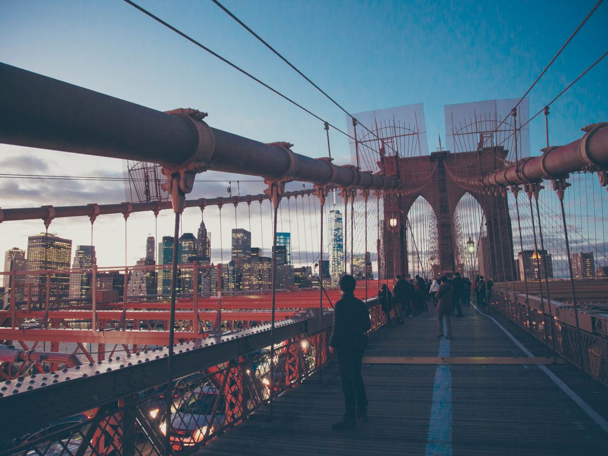 People are walking on a suspension bridge with a city skyline in the background during sunset, featuring prominent red cables.