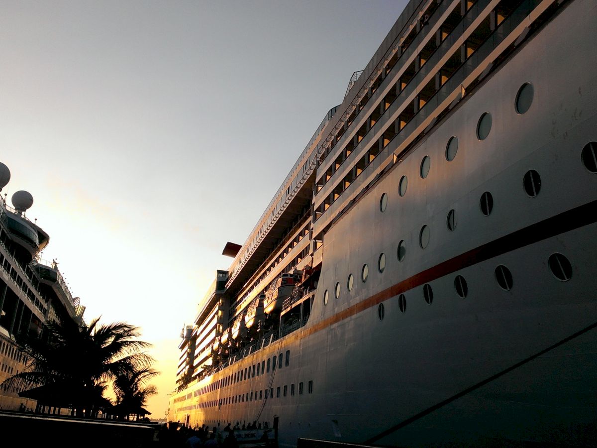 Two large cruise ships are docked side by side at sunset, with palm trees visible between them and the horizon glowing in the background.