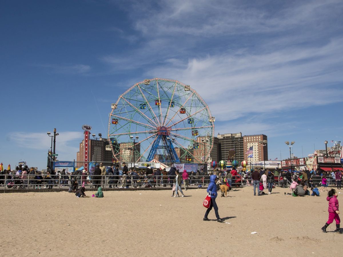 A beach scene with people, a Ferris wheel, and buildings in the background under a partly cloudy sky, creating a lively and colorful atmosphere.