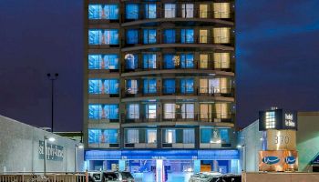 The image shows a multi-story building illuminated at night, with cars parked in front and light trails from passing vehicles on the road.