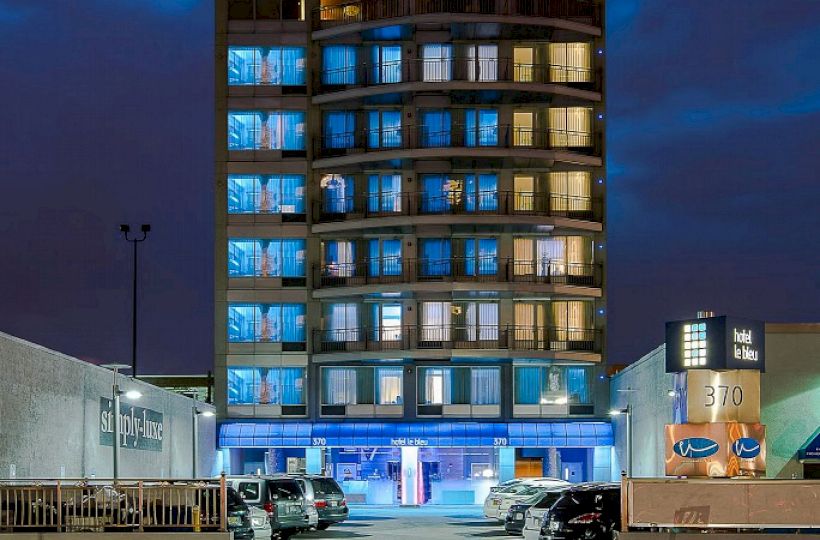 The image shows a multi-story building illuminated at night, with cars parked in front and light trails from passing vehicles on the road.