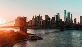 A city skyline at sunset with a bridge spanning a river, towering skyscrapers, and a mixture of natural and artificial light casting soft hues.