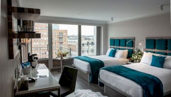 A modern hotel room with two beds, teal accents, a desk, and a city view from large windows.