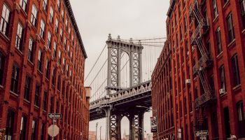 View of a suspension bridge framed between two red brick buildings; the scene has an urban, historic architectural aesthetic, with a moody sky ending the sentence.